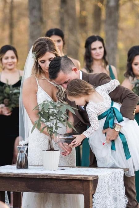 couple holding their child pouring dirt into their tree pot at their wedding ceremony